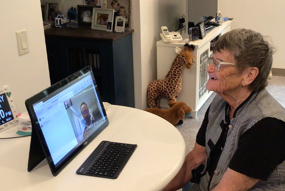 An elderly woman speaking to a caregiver using a tablet placed on a desk with a wireless keyboard in front.