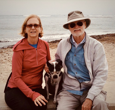 Two older people seated on a bench in front of the beach and ocean with their dog.