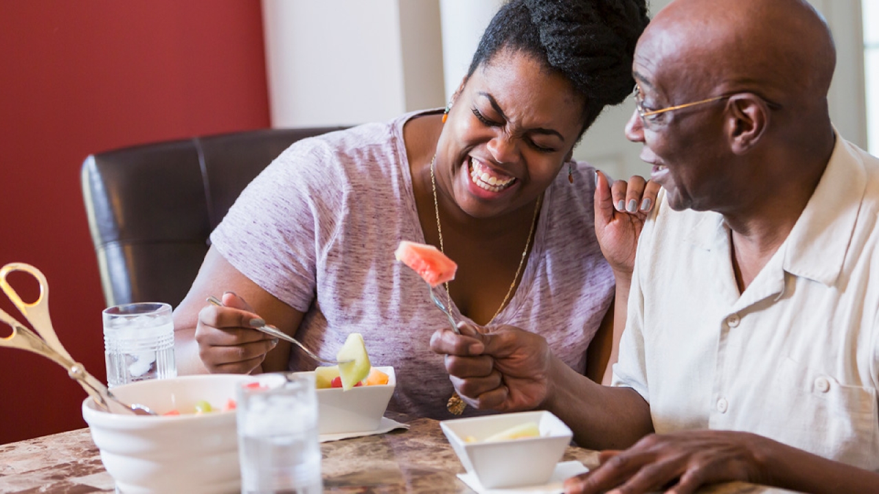Two people laughing in unison while seated at the dining table and eating.