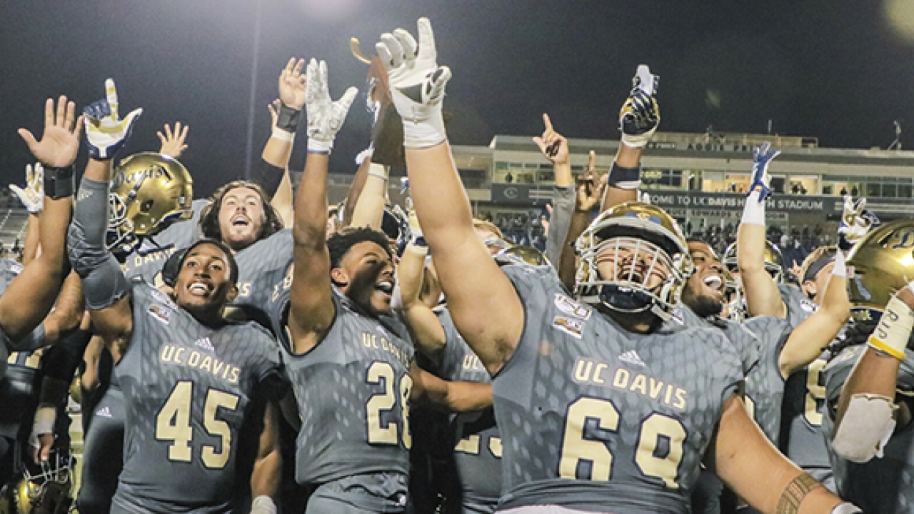 UC Davis football players facing camera in a crowd, hands in the air, outdoors at night.