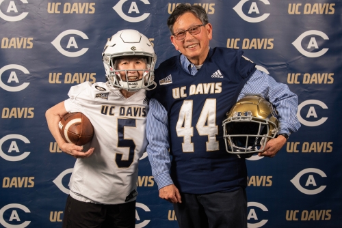 Floyd and Ruth Shimomura pose in football player outfits and gear in front of a dark photo backdrop.