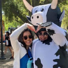 Two people wearing sunglasses pose while forming the shape of a heart on the Quad. One is wearing an inflatable cow costume.